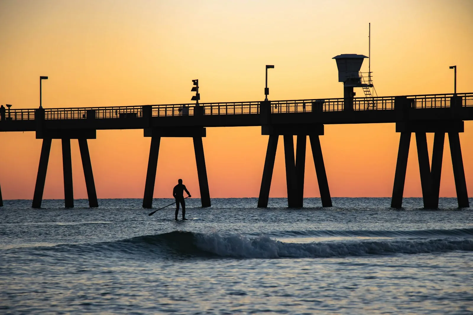 Surfer riding a wave by a pier at sunset